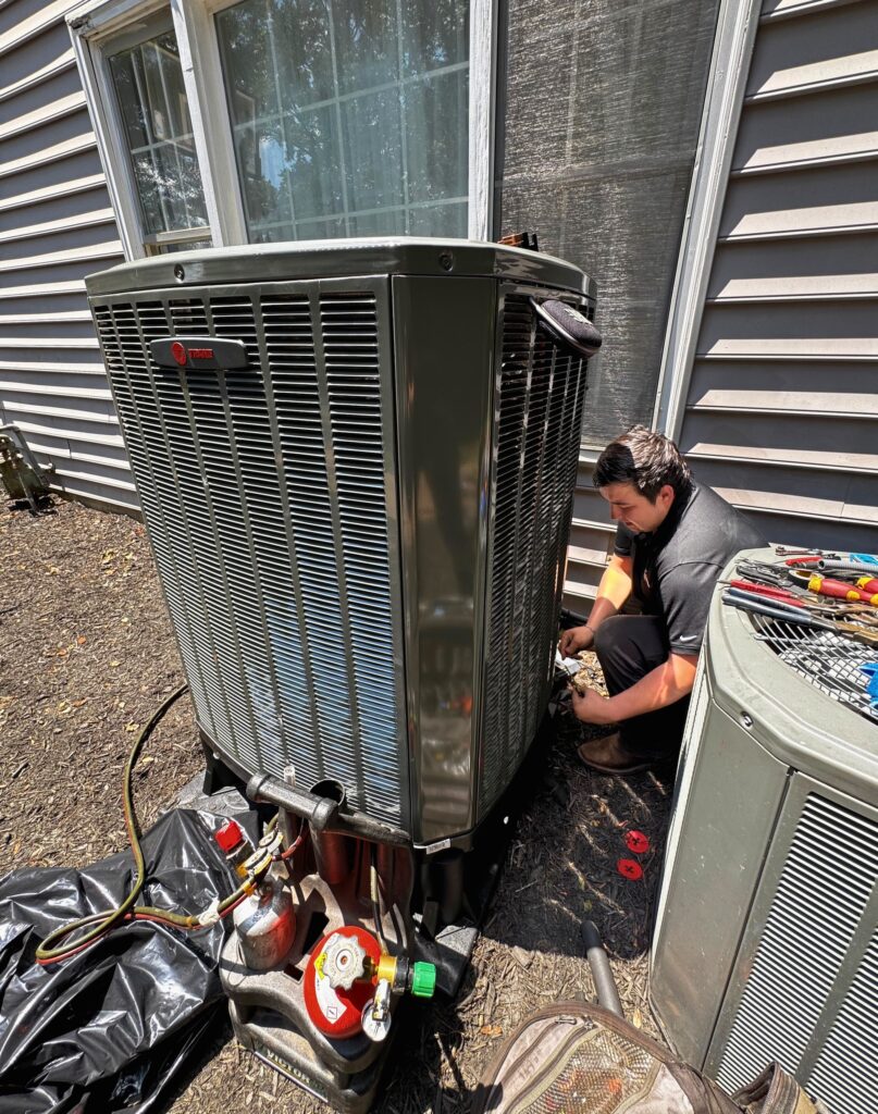 A SwiftPro technician connecting a new Trane outdoor air conditioning unit beside a Northern Virginia house, with refrigerant tanks and the old condenser next to it
