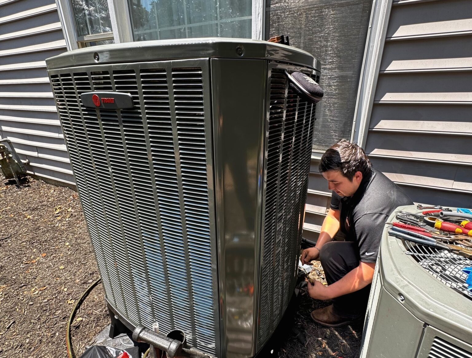 A SwiftPro technician kneeling beside a new Trane outdoor air conditioning unit at a Northern Virginia house, with tools laid out on the old condenser