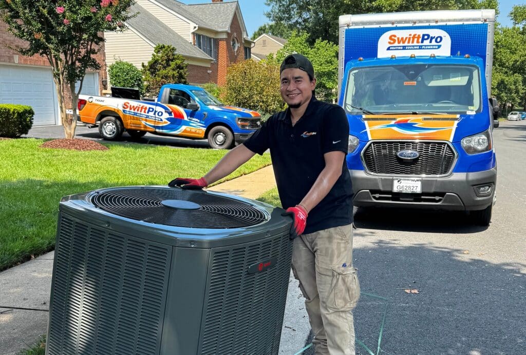 A smiling SwiftPro technician in a black company polo and red work gloves beside a new Trane outdoor air conditioning unit on a Northern Virginia street, a wrapped SwiftPro pickup and box truck behind him