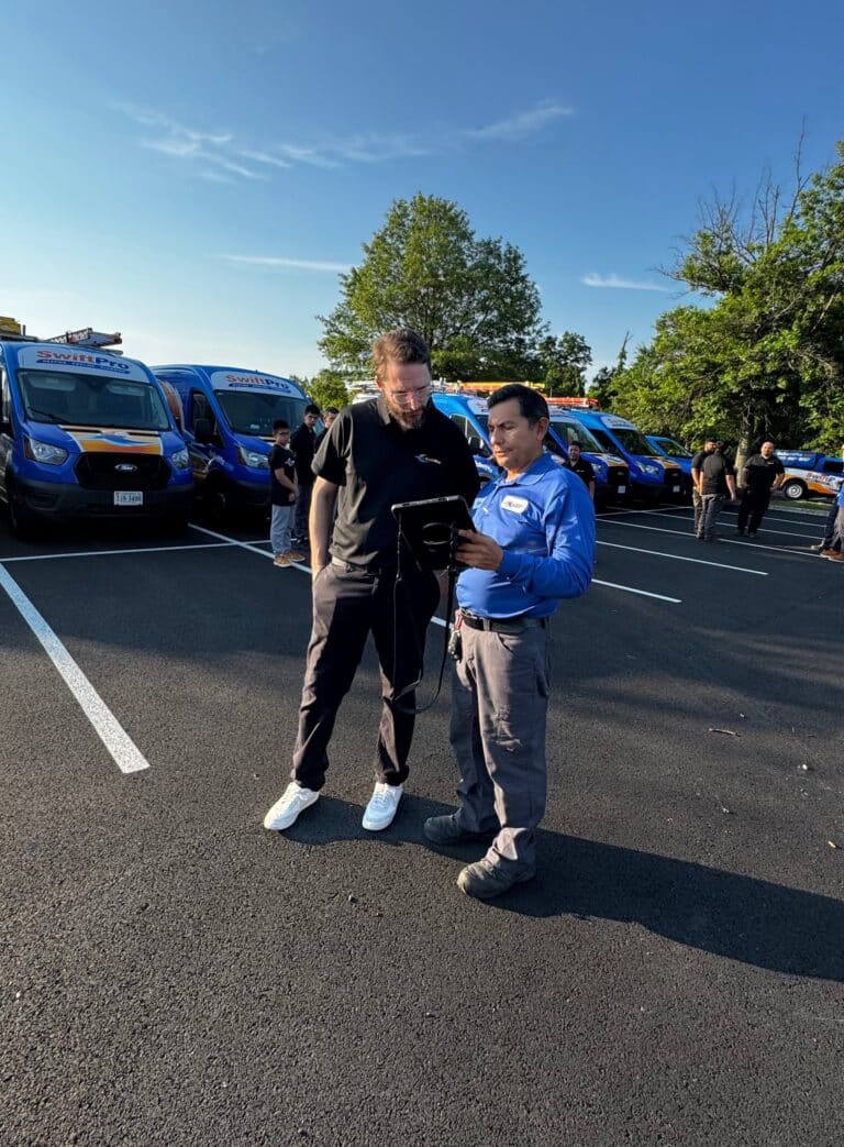 Two SwiftPro technicians, one in a blue company polo and one in black, going over a work order on a tablet in front of the wrapped SwiftPro van fleet in Northern Virginia
