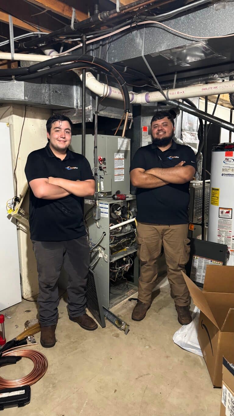 Two SwiftPro technicians in black company polos standing with their arms crossed beside a gas furnace and a water heater in an unfinished Northern Virginia basement