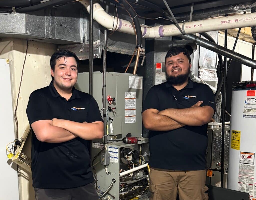 Two SwiftPro HVAC technicians in black company polos standing arms crossed in a Northern Virginia basement mechanical room, in front of a gas furnace with its front panel off and a water heater beside it