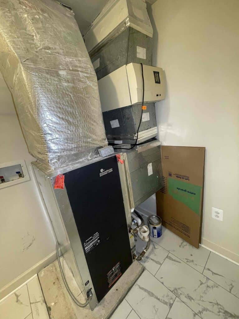 A black ClimateMaster Tranquility 16 water source heat pump on a pad in a small Northern Virginia utility room, two stainless braided water hoses and a white PVC condensate trap at its lower right and a humidifier on the foil-wrapped supply duct above it