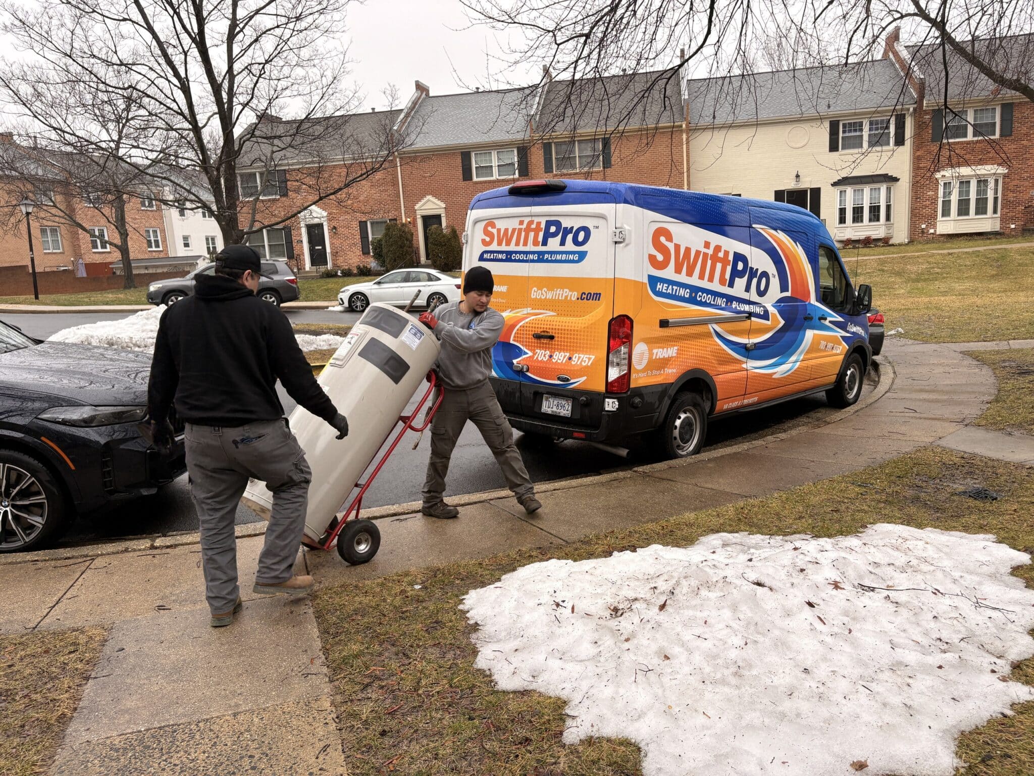 Two SwiftPro plumbers wheeling a new water heater up a Northern Virginia walkway on a hand truck, beside the wrapped van