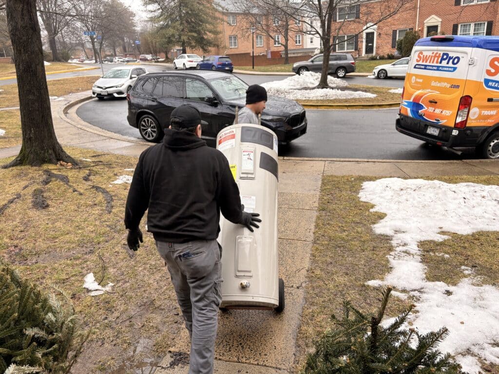 A SwiftPro technician carrying a new water heater across a driveway to a Northern Virginia home, with the SwiftPro service van parked at the curb