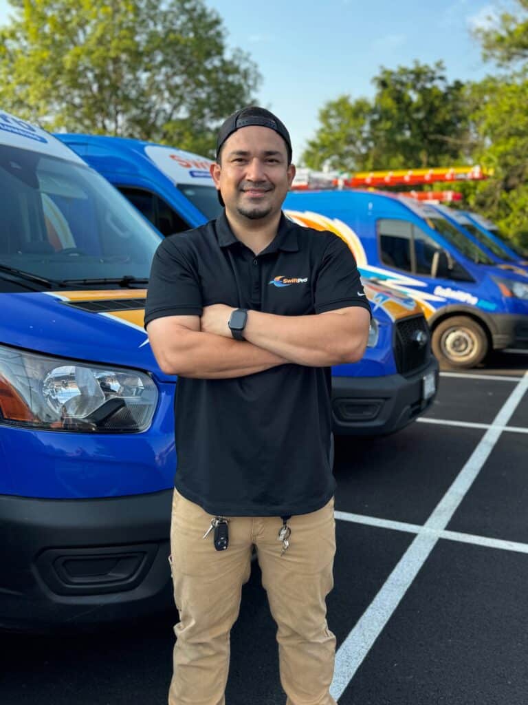 A smiling SwiftPro HVAC technician in a black company polo and a backward cap, arms crossed, in front of a row of wrapped SwiftPro service vans in Northern Virginia