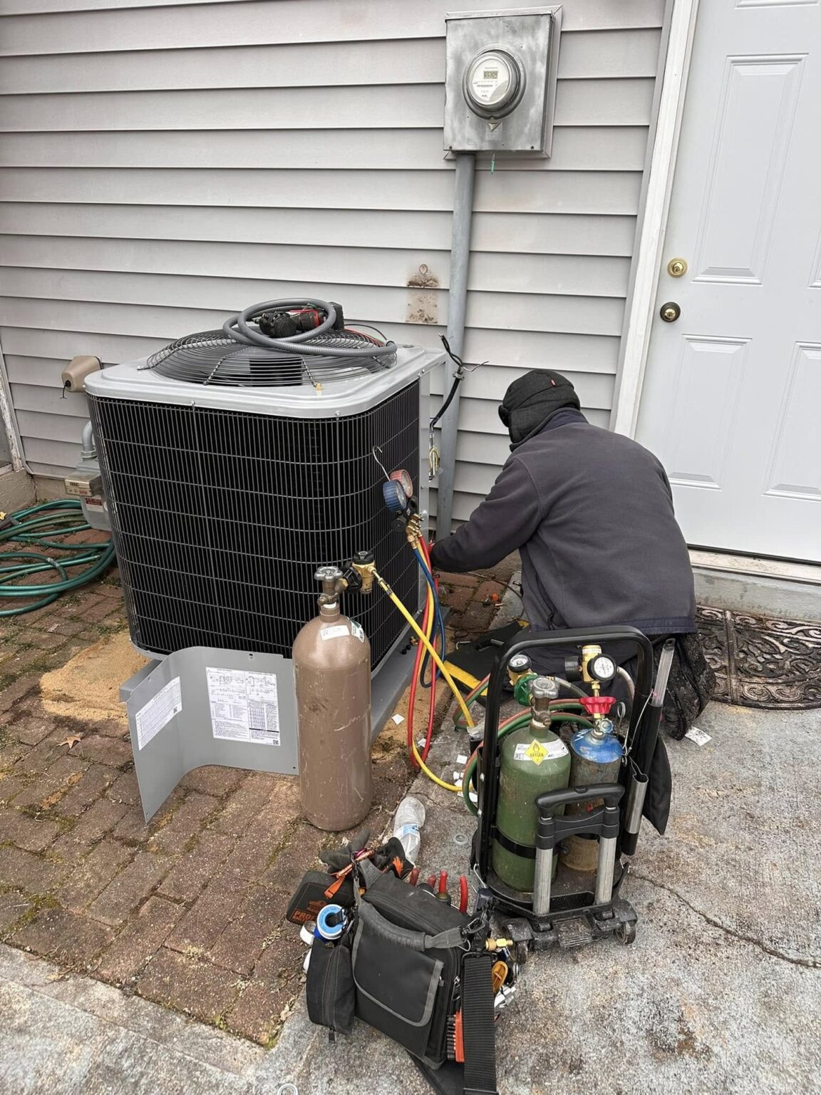 A SwiftPro technician kneeling at an outdoor condenser with refrigerant recovery tanks and gauges