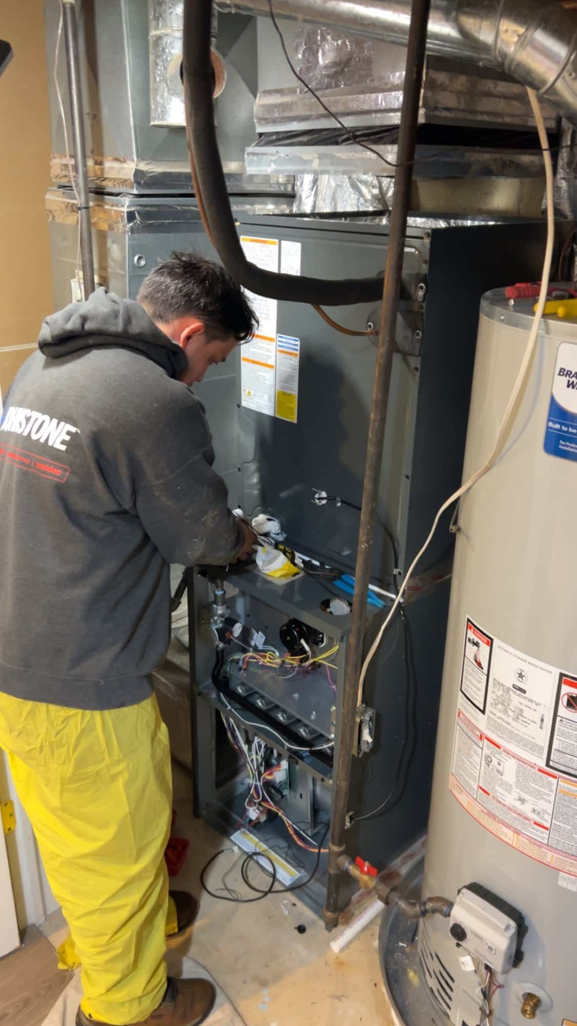 A technician working inside an open furnace cabinet in a Northern Virginia utility closet, beside a Bradford White gas water heater