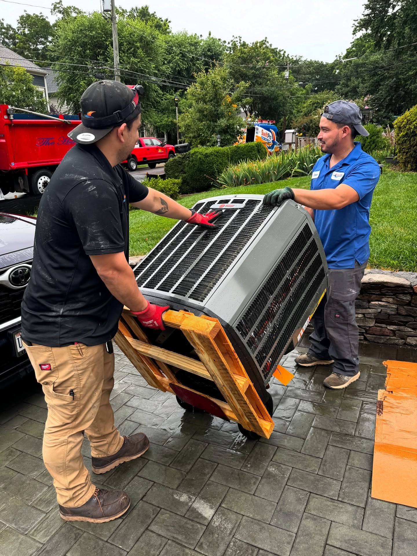 Two SwiftPro technicians wheeling a new Trane condenser up a driveway on an AC replacement in Ashburn, VA, wrapped SwiftPro van behind them