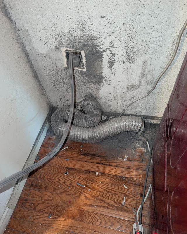 Behind a dryer in Ashburn, Virginia, before a SwiftPro dryer vent cleaning: grey lint up the wall around the vent opening and a crushed foil hose on the floor