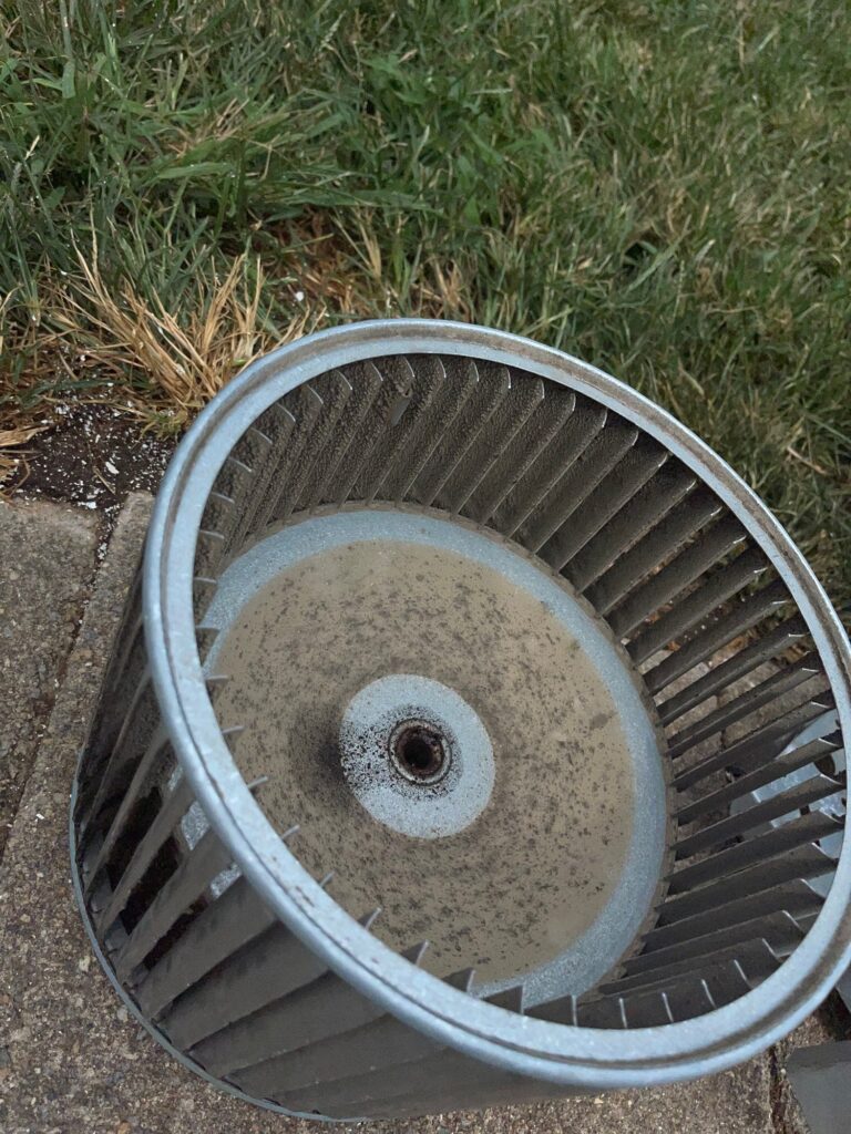 A furnace blower wheel pulled out on a SwiftPro heating job in Northern Virginia, set on the grass before cleaning, dust caked across its back plate and blades