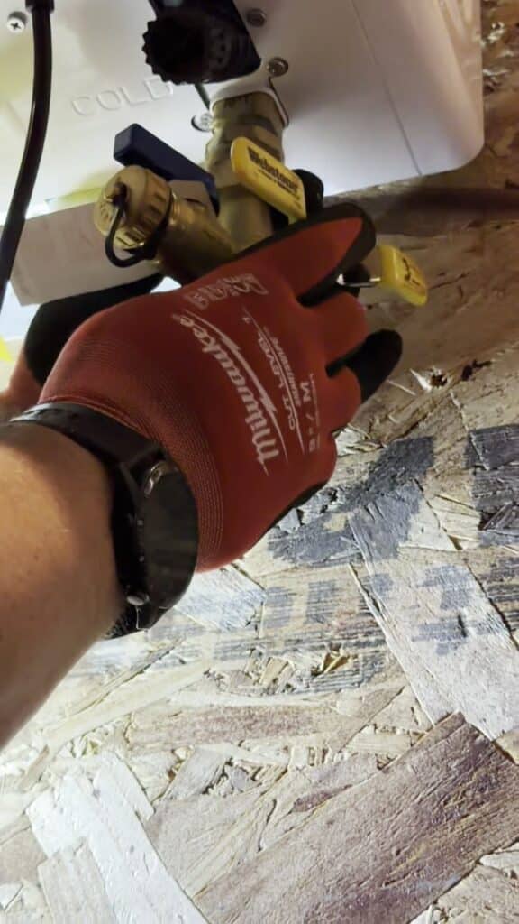 A SwiftPro plumber's gloved hand on the brass service valves with yellow handles under a new tankless water heater in Northern Virginia, the ports a descaling pump connects to