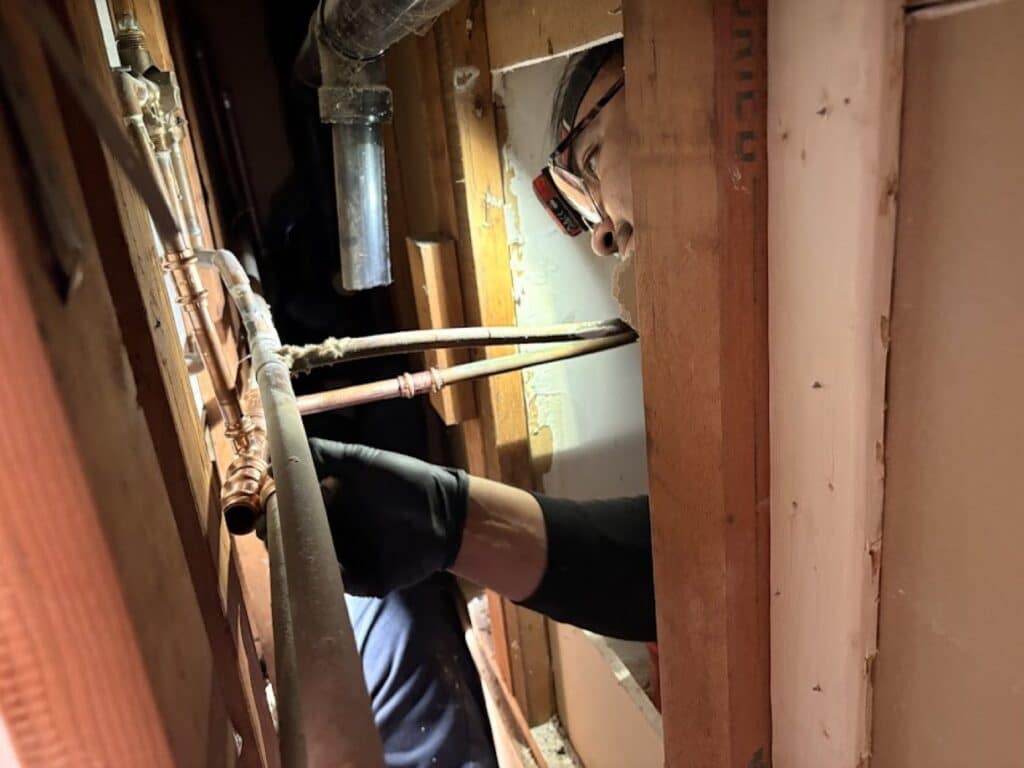 A SwiftPro plumber in safety glasses and black gloves reaching through an opened wall to repair copper water lines at a home in Reston, Virginia