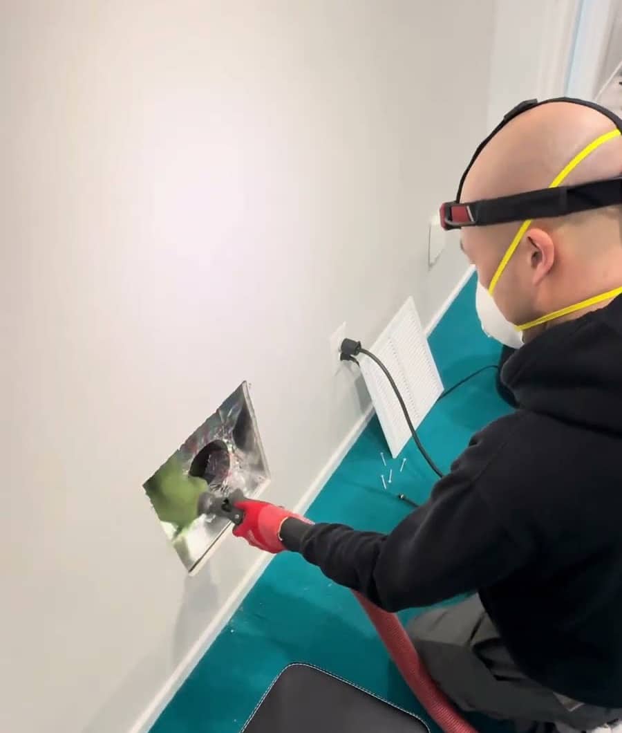 A SwiftPro technician in a mask and headlamp running a cleaning tool into an open wall register in a Northern Virginia home, a drop cloth on the floor