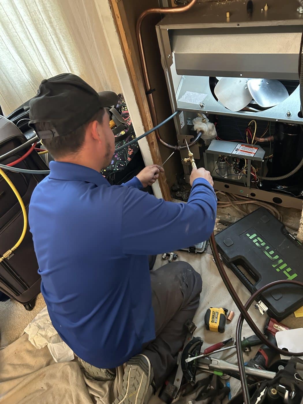 A SwiftPro technician in a blue polo kneeling at an open HVAC cabinet, working on the refrigerant piping with his tools on a drop cloth