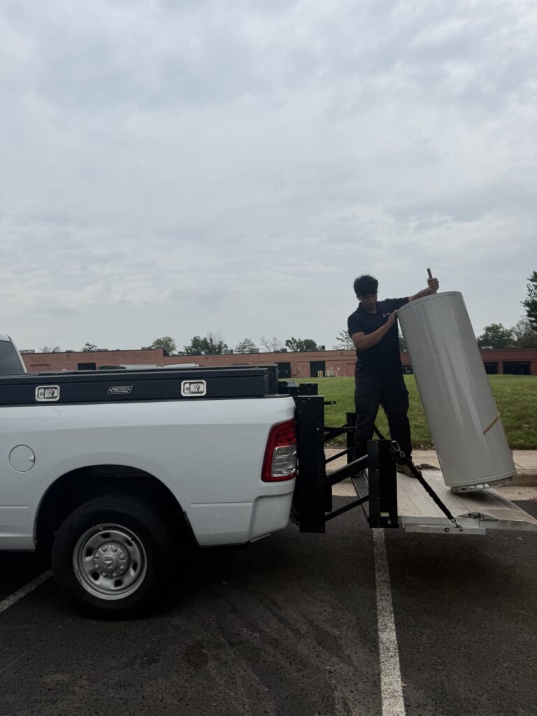 A SwiftPro plumber in a black company polo lowering a new tank water heater off a pickup truck's liftgate in a Northern Virginia parking lot
