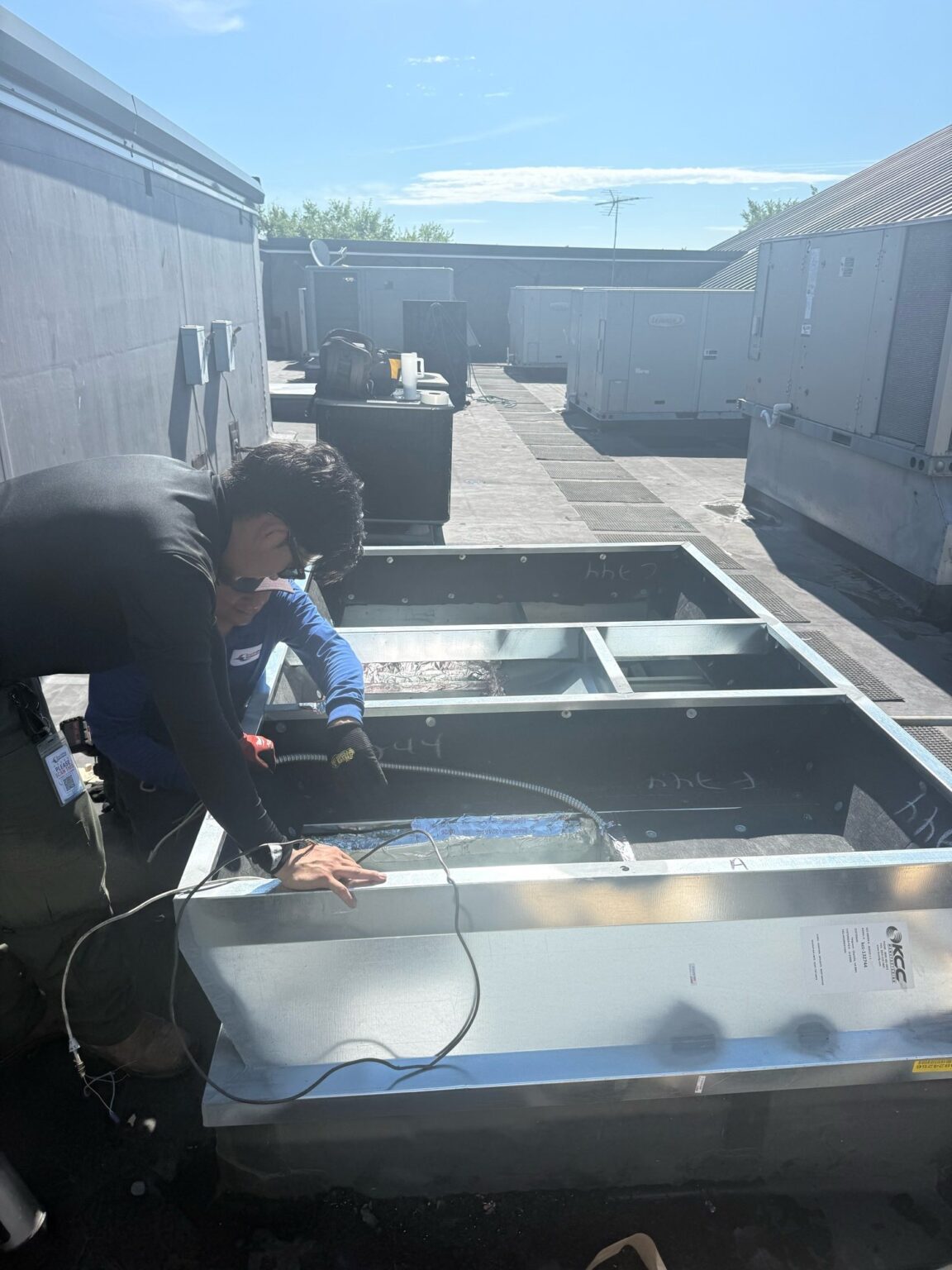 Two SwiftPro technicians at work on a new rooftop unit curb adapter on a flat commercial roof in Northern Virginia, with a row of packaged rooftop HVAC units behind them
