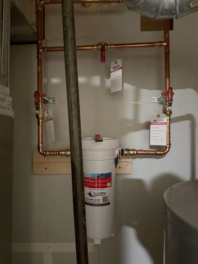 A main water line in a Northern Virginia basement after a SwiftPro whole-house filter install: a copper bypass loop with red-handled ball valves, each one tagged for valve inspection
