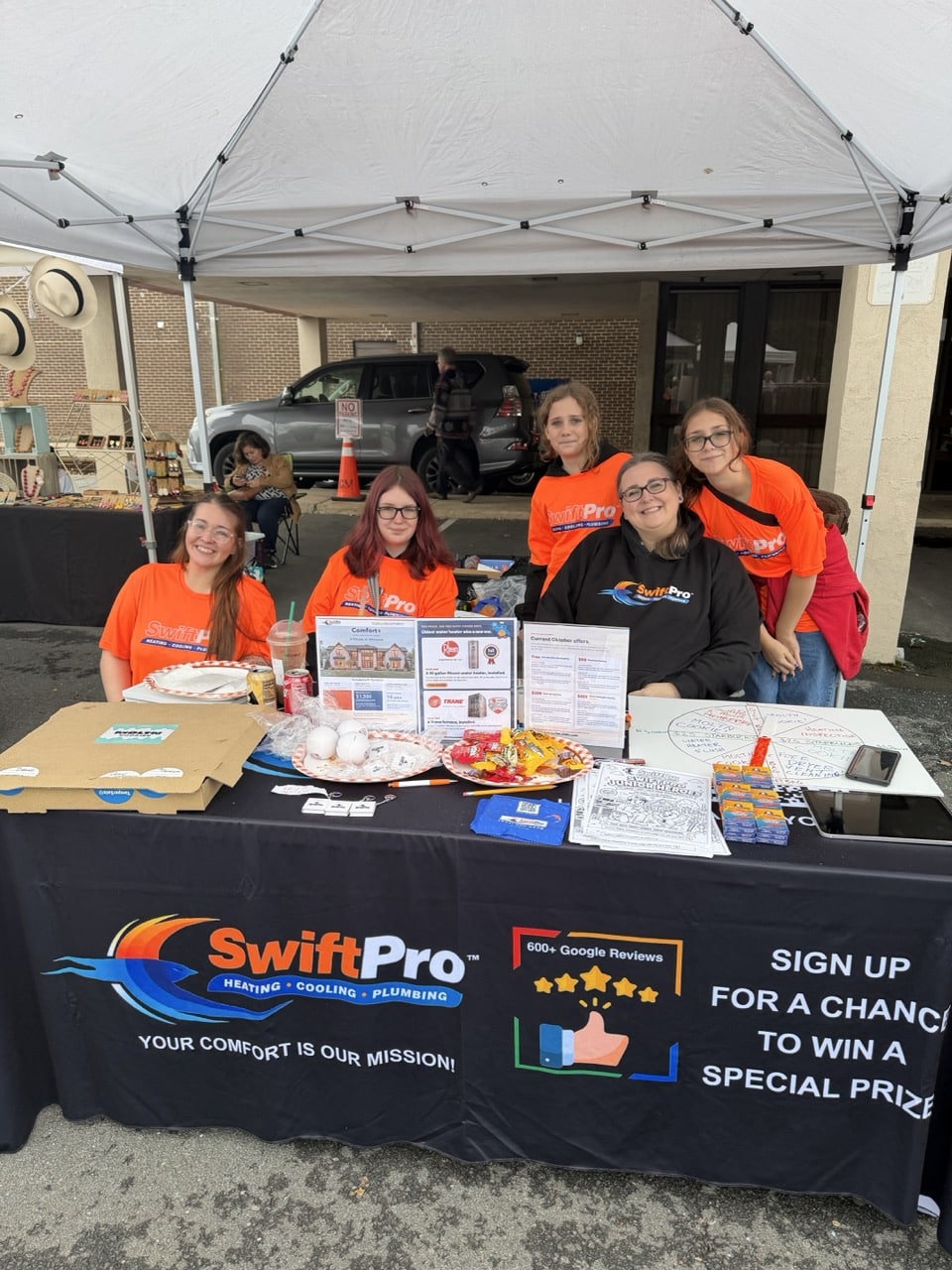 Five SwiftPro team members in orange and black SwiftPro shirts at the SwiftPro booth at the Vienna Oktoberfest in Vienna, Virginia, with Comfort+ membership flyers on the table