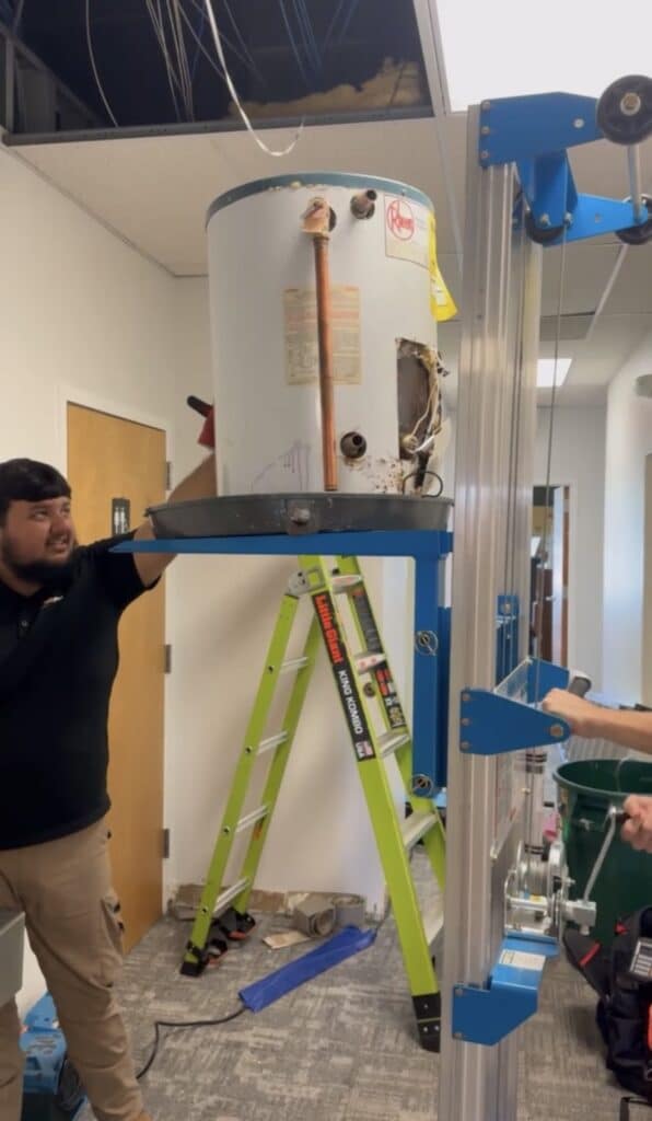 A SwiftPro plumber lifting an old Rheem water heater out of a Northern Virginia building with a hoist and a drain pan, on its way to replacement
