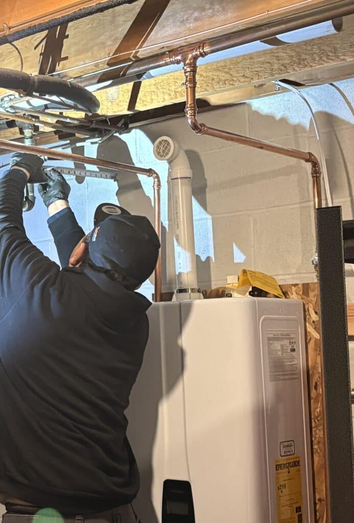 A SwiftPro plumber in a cap and work gloves fitting new copper water lines overhead in a Northern Virginia basement, a new white condensing tankless water heater below him with its white PVC venting on top