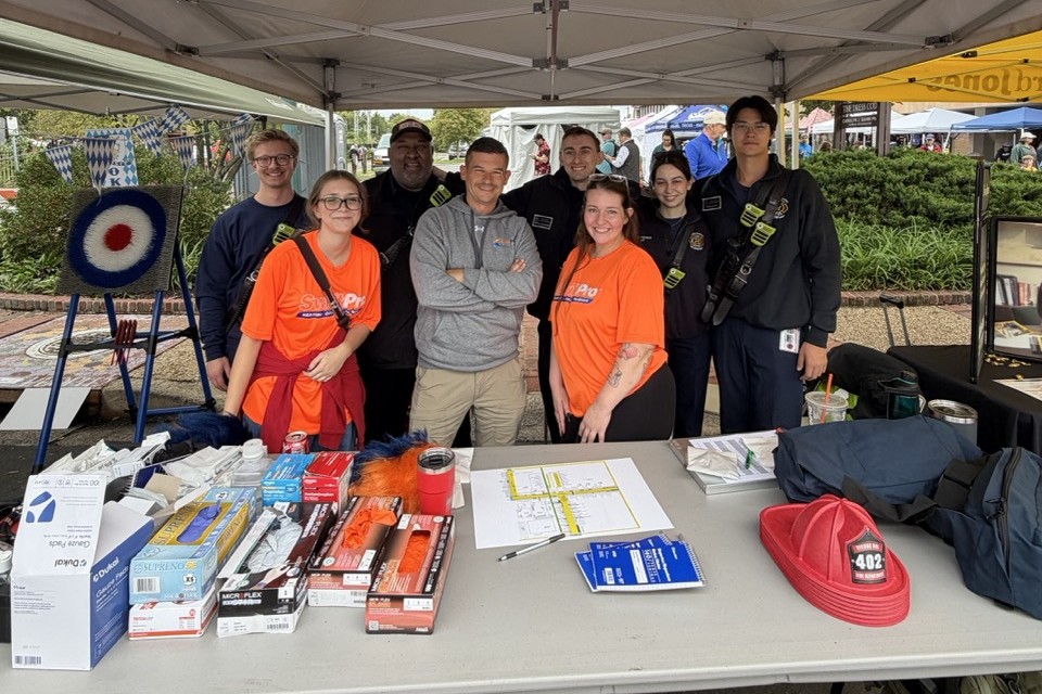 SwiftPro owner Tyler Griffin, center, with SwiftPro team members in orange shirts and local firefighters behind a fire safety table at the Vienna Oktoberfest in Vienna, Virginia