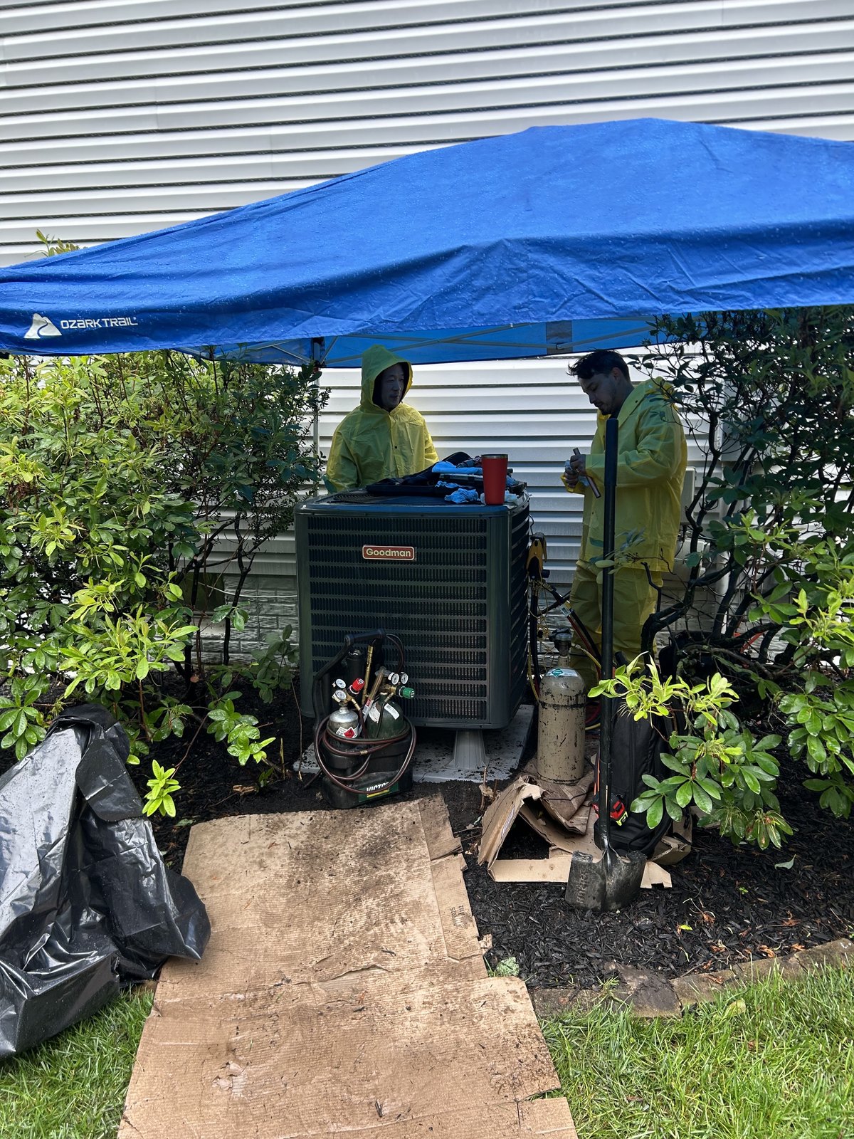 Two SwiftPro HVAC technicians in rain gear working on a Goodman condenser under a pop-up canopy at a Northern Virginia house, brazing tanks beside them