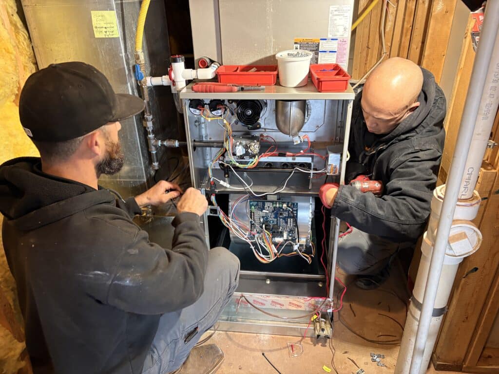 Two SwiftPro technicians wiring the control board of a new Carrier gas furnace in an unfinished basement in Ashburn, Virginia, a red parts tray on top of the cabinet
