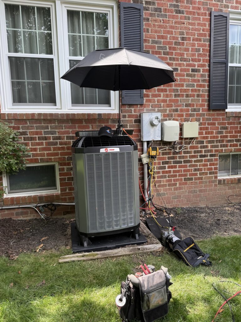 A new Trane outdoor unit being installed on a pad beside a brick house in Northern Virginia, an umbrella rigged over it, hoses run to the wall and tool bags on the grass