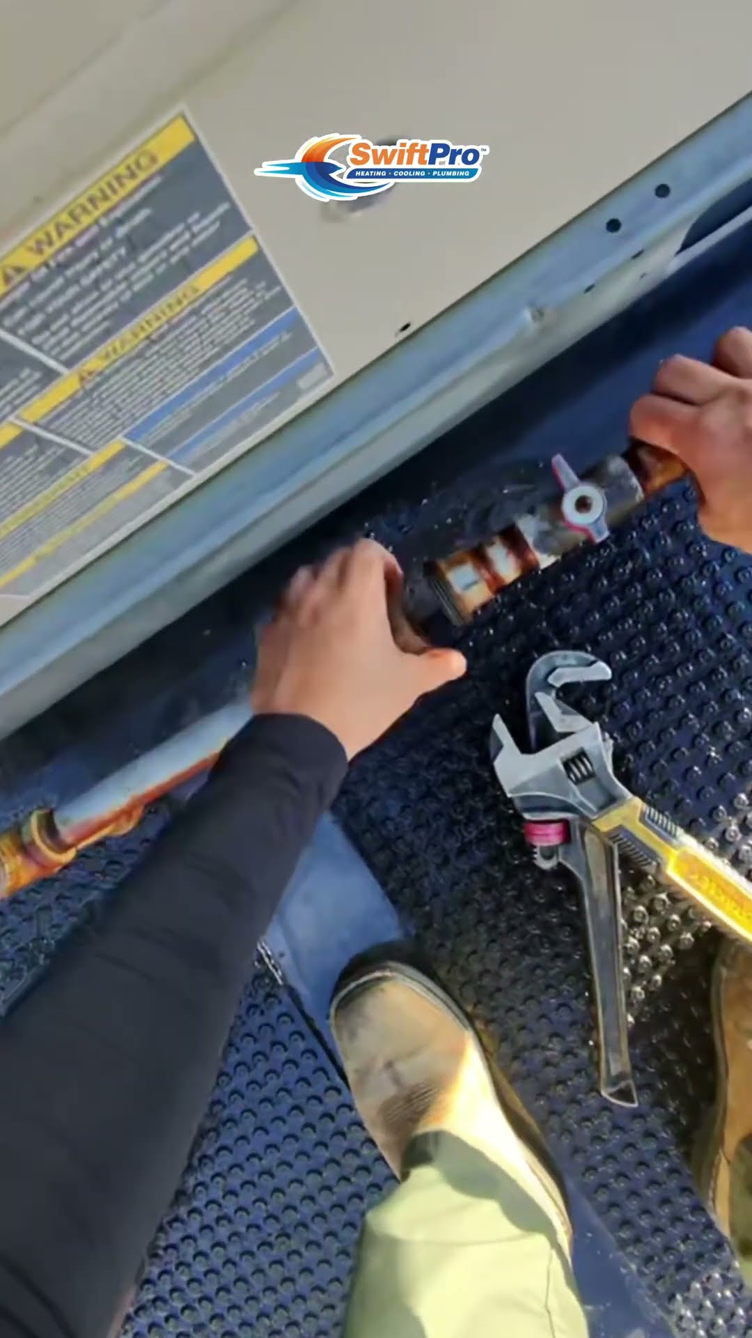 First-person rooftop AC replacement on a wet commercial roof: a hand turns a gas valve at a package unit with an adjustable wrench nearby, a tool bag and hoses sit on a walkway pad, and the new roofto
