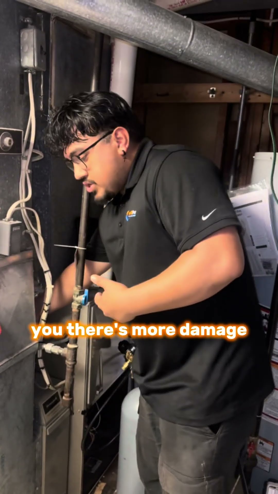 A tech in a black SwiftPro polo and glasses stands in an unfinished basement by the furnace and AC coil, explaining how a condensate overflow safety switch shuts the system off before a clogged drain 