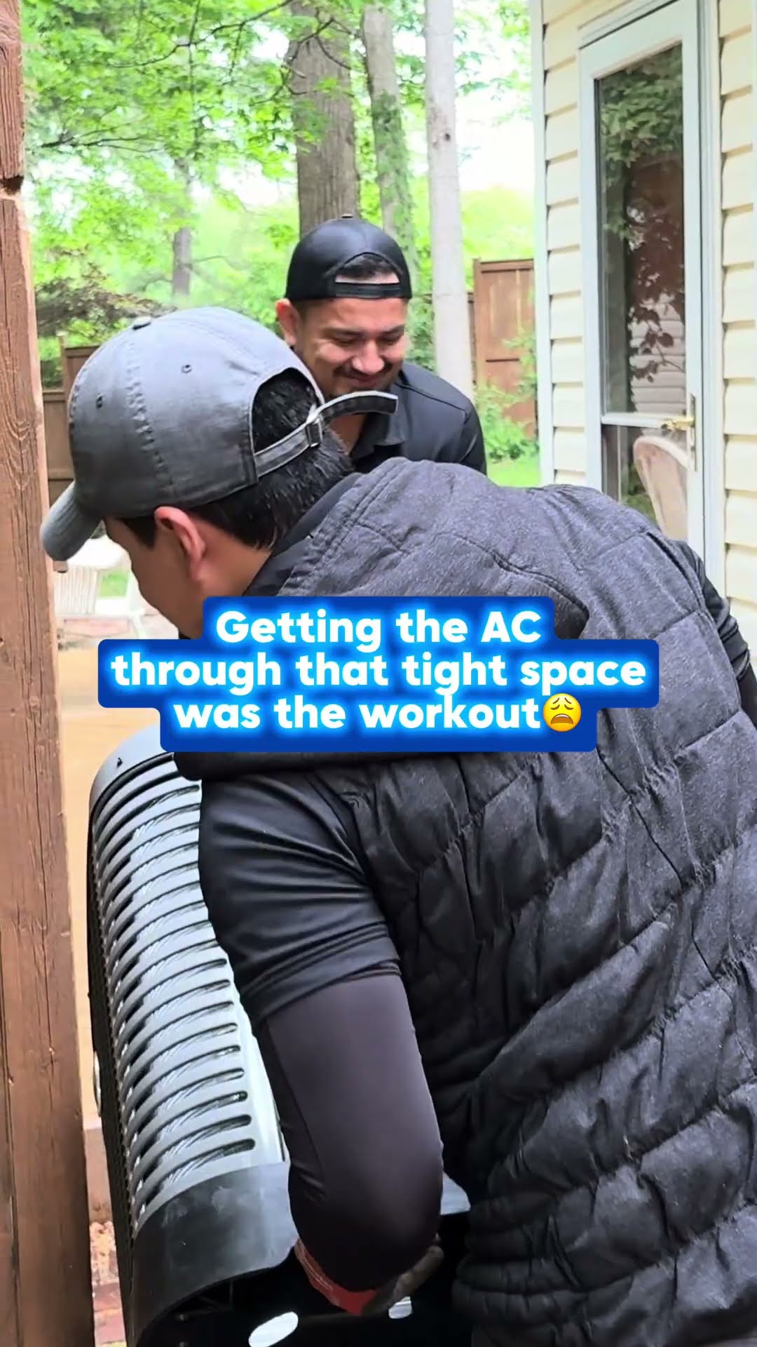 Three techs squeeze a new AC condenser through a narrow wooden side-yard gate beside a house, one smiling while lifting.