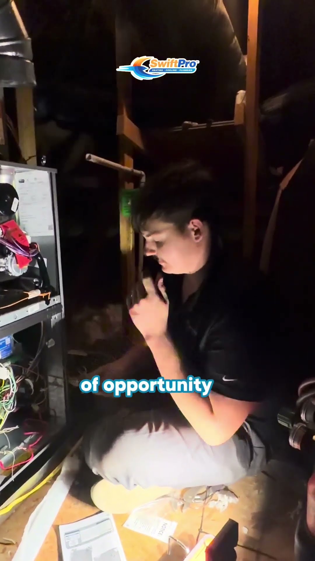 A young tech in a black polo sits cross-legged in an attic beside an open furnace with wiring and manuals, talking to camera about opportunity and instruction in the trades.