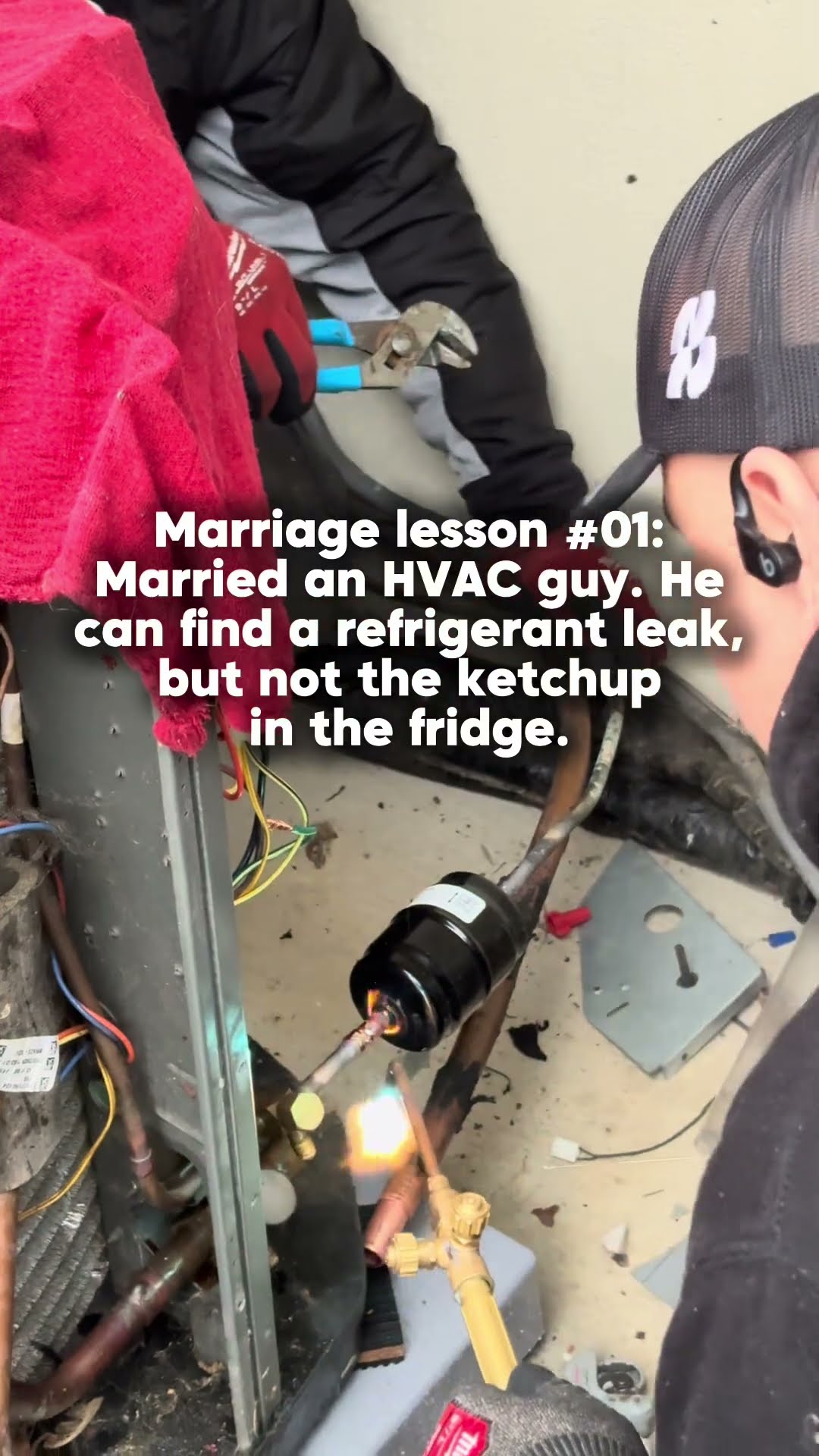 Two techs at an outdoor AC condenser with the panel off, one brazing a filter drier into the refrigerant line with a torch.