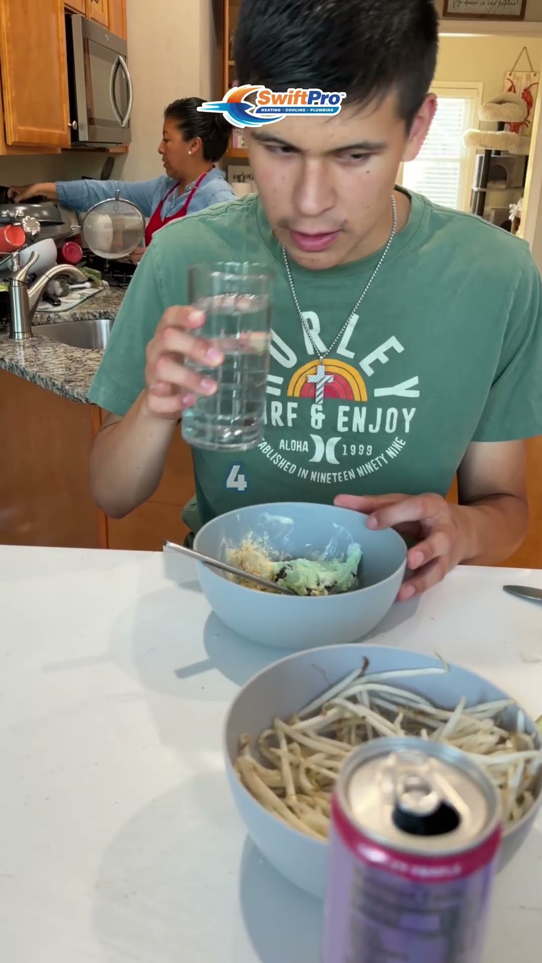 In a family kitchen, a teen in a green T-shirt sips tap water and reacts while a woman cooks in the background.