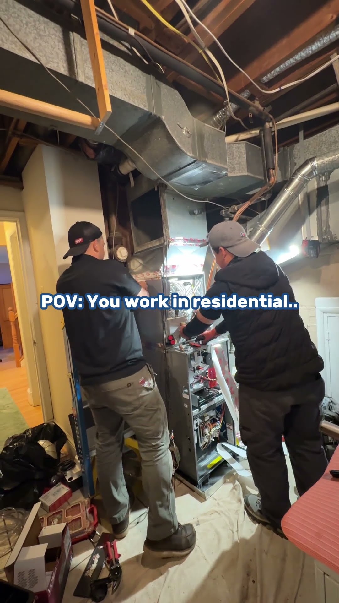 Two techs in black jackets and caps work on an open gas furnace in an unfinished basement under grey ductwork, with tool bags and a drop cloth on the floor.