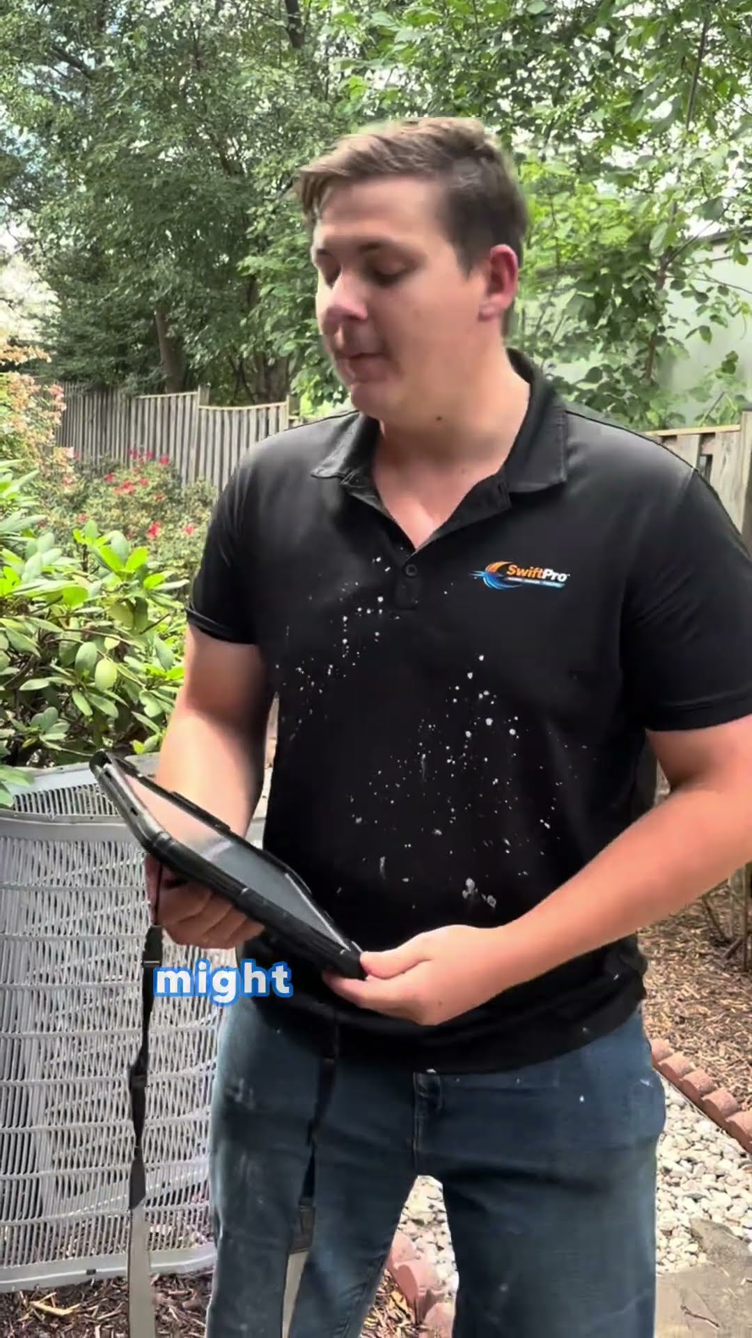 A young tech in a black SwiftPro polo stands beside an AC condenser in a backyard holding a tablet and explains how dirty AC coils raise costs on a 15-year-old unit.