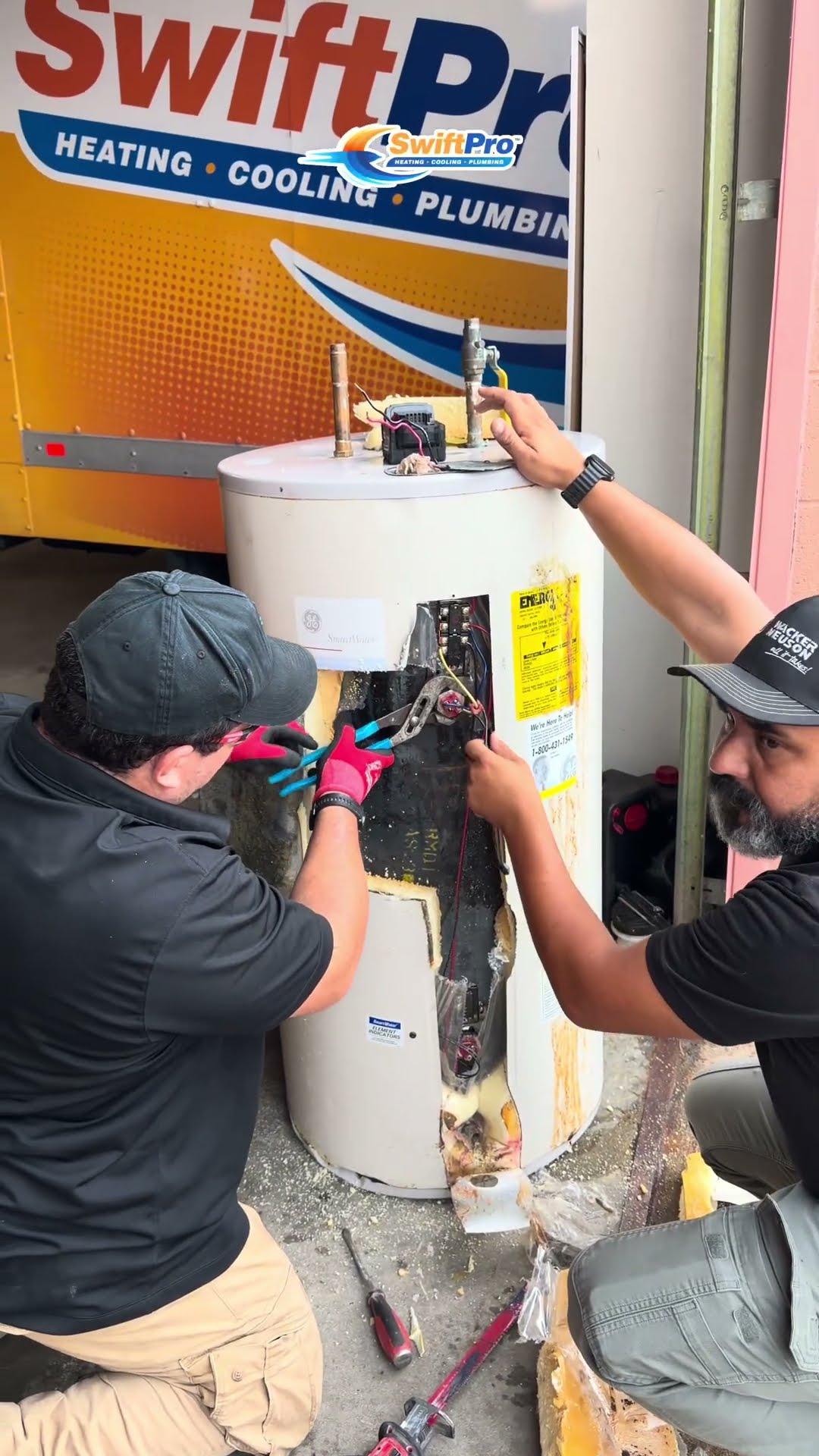 Two techs cut open an old GE electric water heater in front of a SwiftPro van at the shop, peeling back the insulation to show the inside of the tank.