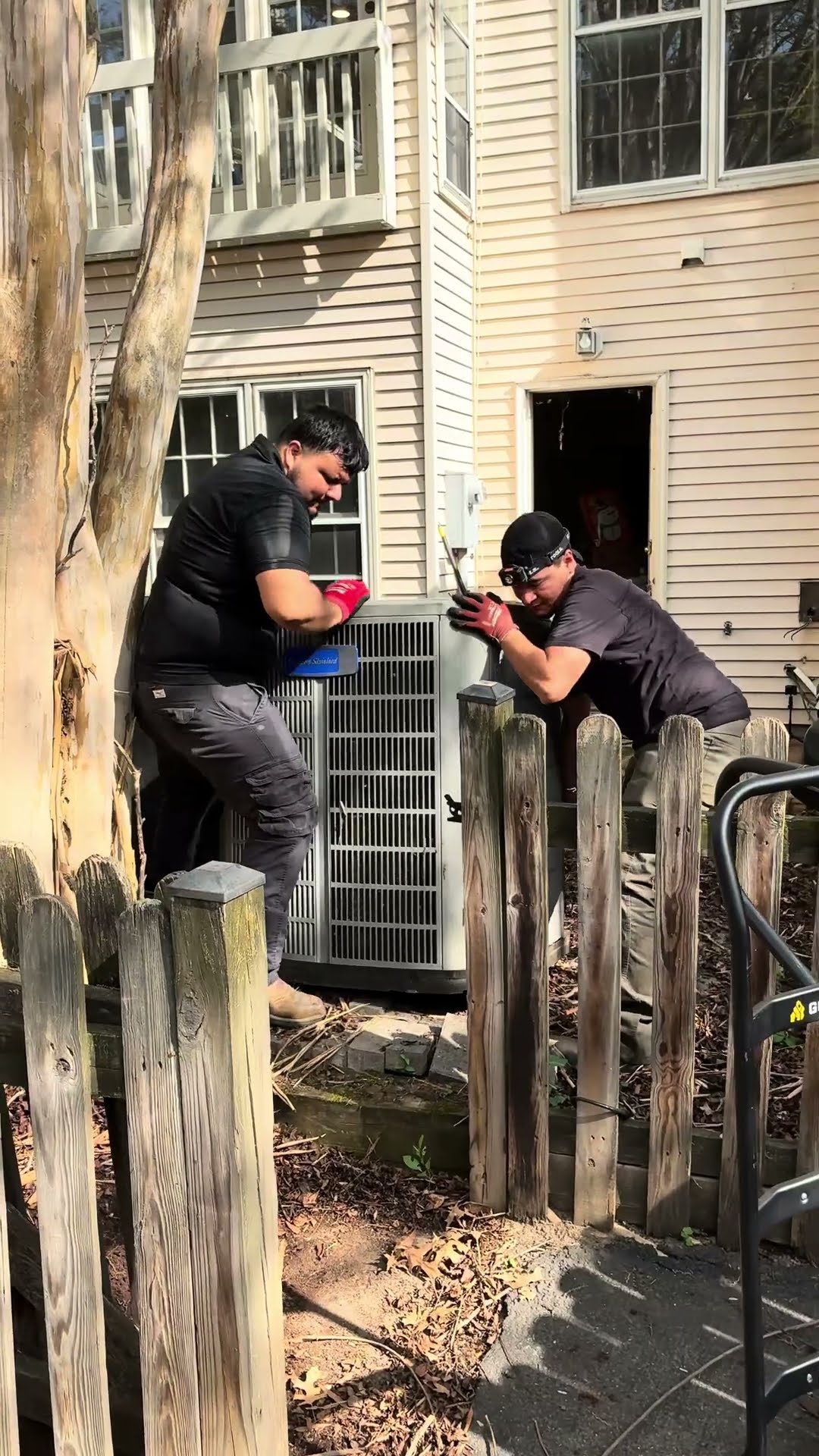 Two techs lift a new AC condenser into place behind a townhouse, beside a wooden fence.