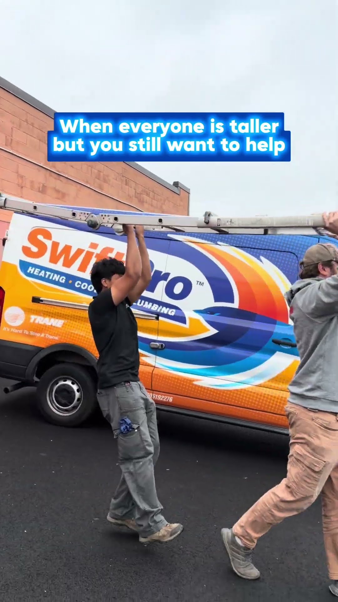 Crew members carry an extension ladder overhead past wrapped SwiftPro vans (Trane badge and VA CLASS A license number on the van) in the shop lot; the shortest one jumps to reach it.