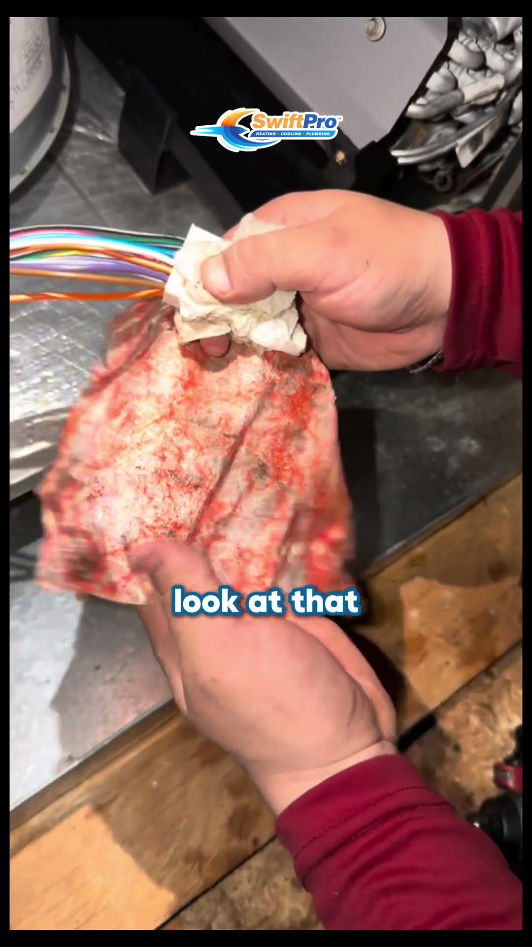 Hands in maroon sleeves wipe rust-red dust off a bundle of control wires with a rag inside an air handler or furnace cabinet.