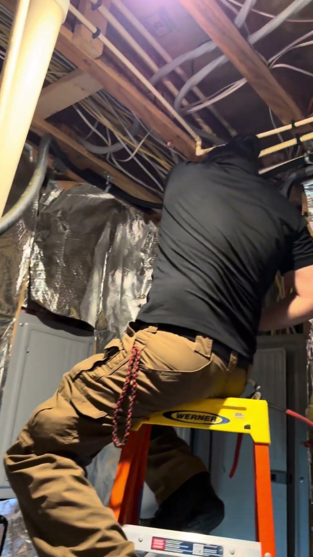 A tech on a Werner ladder runs refrigerant line and wiring through the joists of an unfinished basement ceiling lined with foil-faced insulation, using a cordless drill.