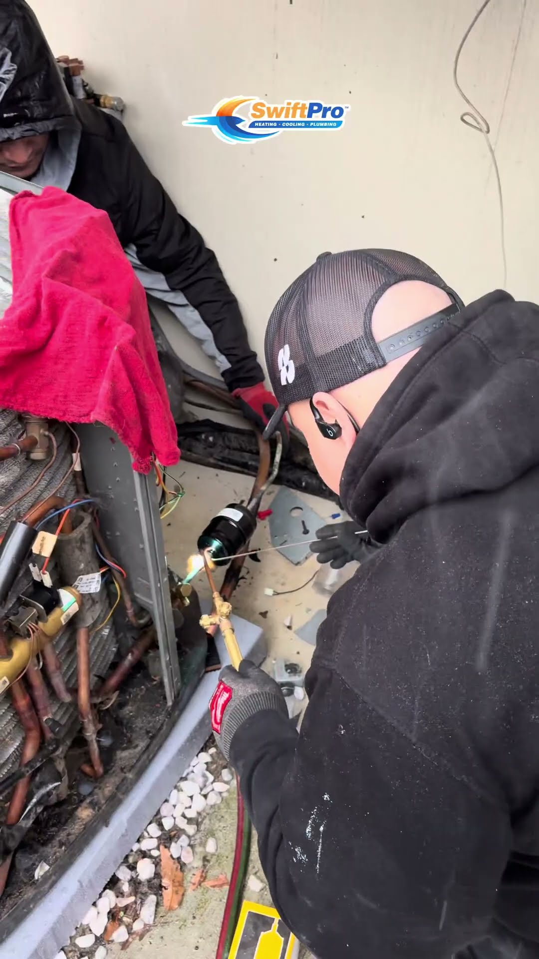 A tech in a backward black cap and hoodie brazes copper at an opened condenser with a red rag over the coil, another tech handles a pink refrigerant recovery cylinder by an AC condenser beside a house