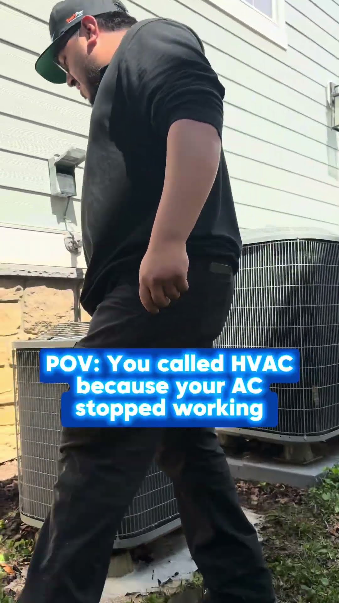 A tech in a black t-shirt and cap walks across a backyard lawn toward the camera, then strides past two AC condensers beside a house.