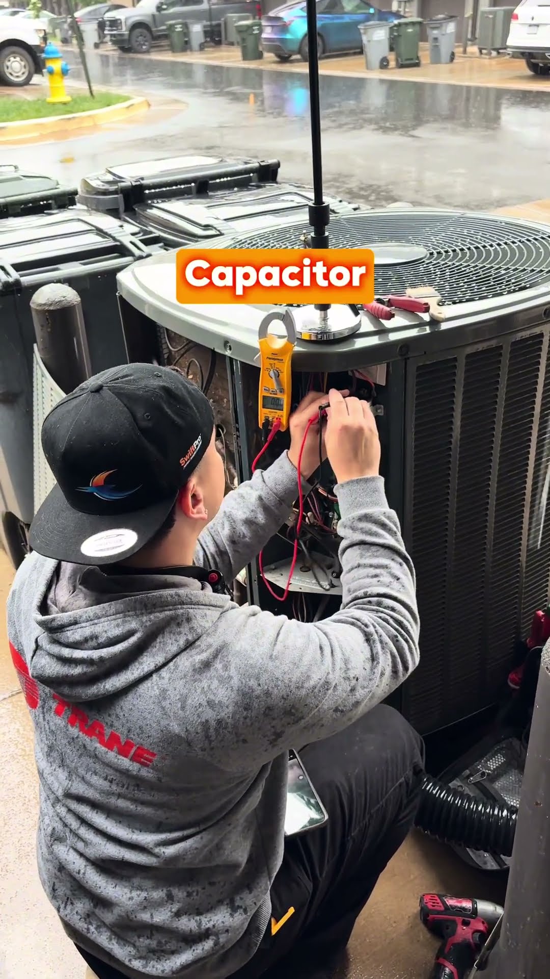 A tech in a gray Trane hoodie and black SwiftPro cap tests a condenser capacitor with a clamp meter at a row of AC condensers outside townhomes in the rain.