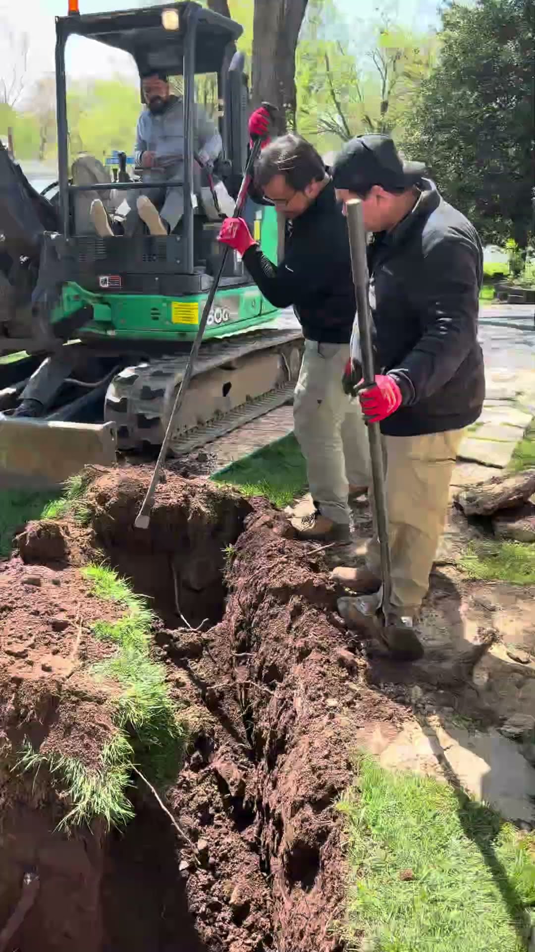 Sewer line repair: a tech with a RIDGID drain camera kneels by a house, a mini excavator digs a trench across a front yard while techs work it with shovels and a bar, and a tech in the trench beside t