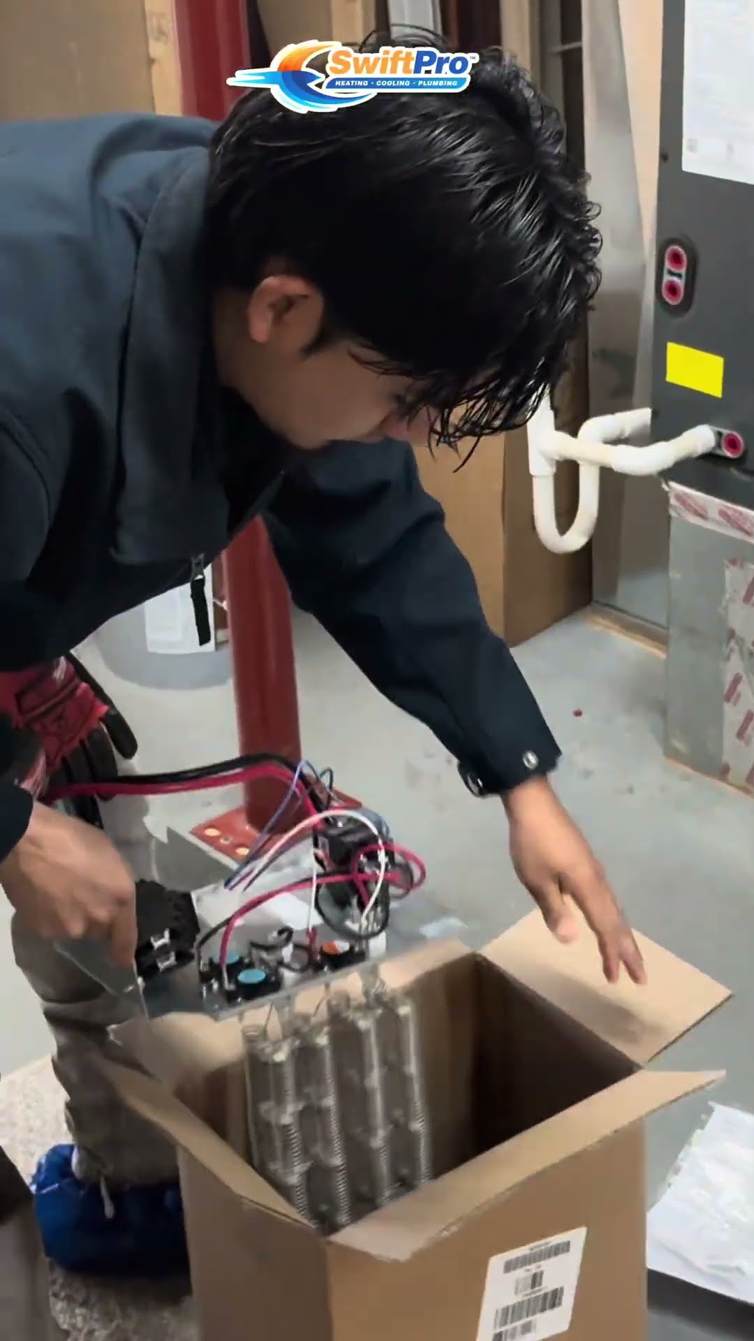 A young tech in a navy work jacket wires a new electric heat strip kit into an open air handler in a utility room, using a Milwaukee tool, with the boxed heating elements on the floor.
