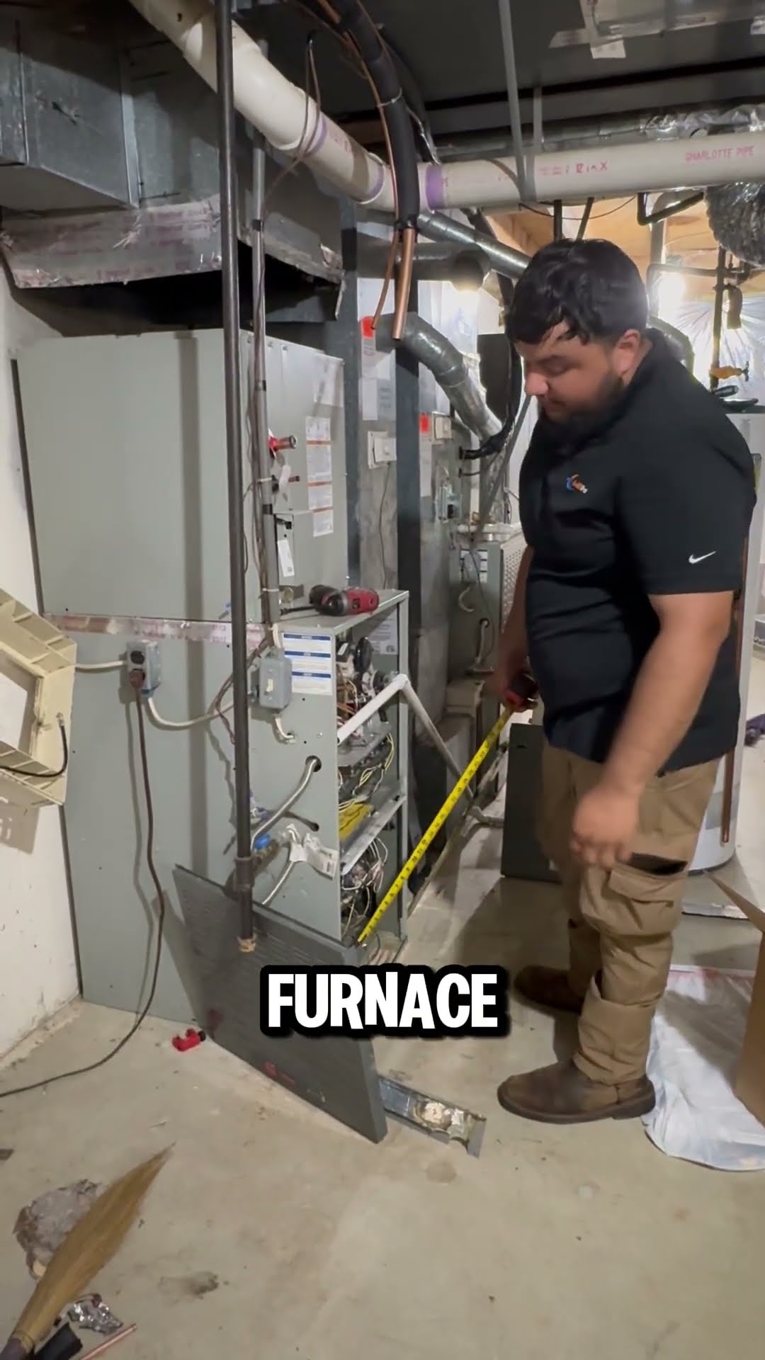 A bearded tech in a black SwiftPro polo and tan cargo pants measures an old furnace with a tape measure in an unfinished basement, ahead of installing a new AC coil and system.