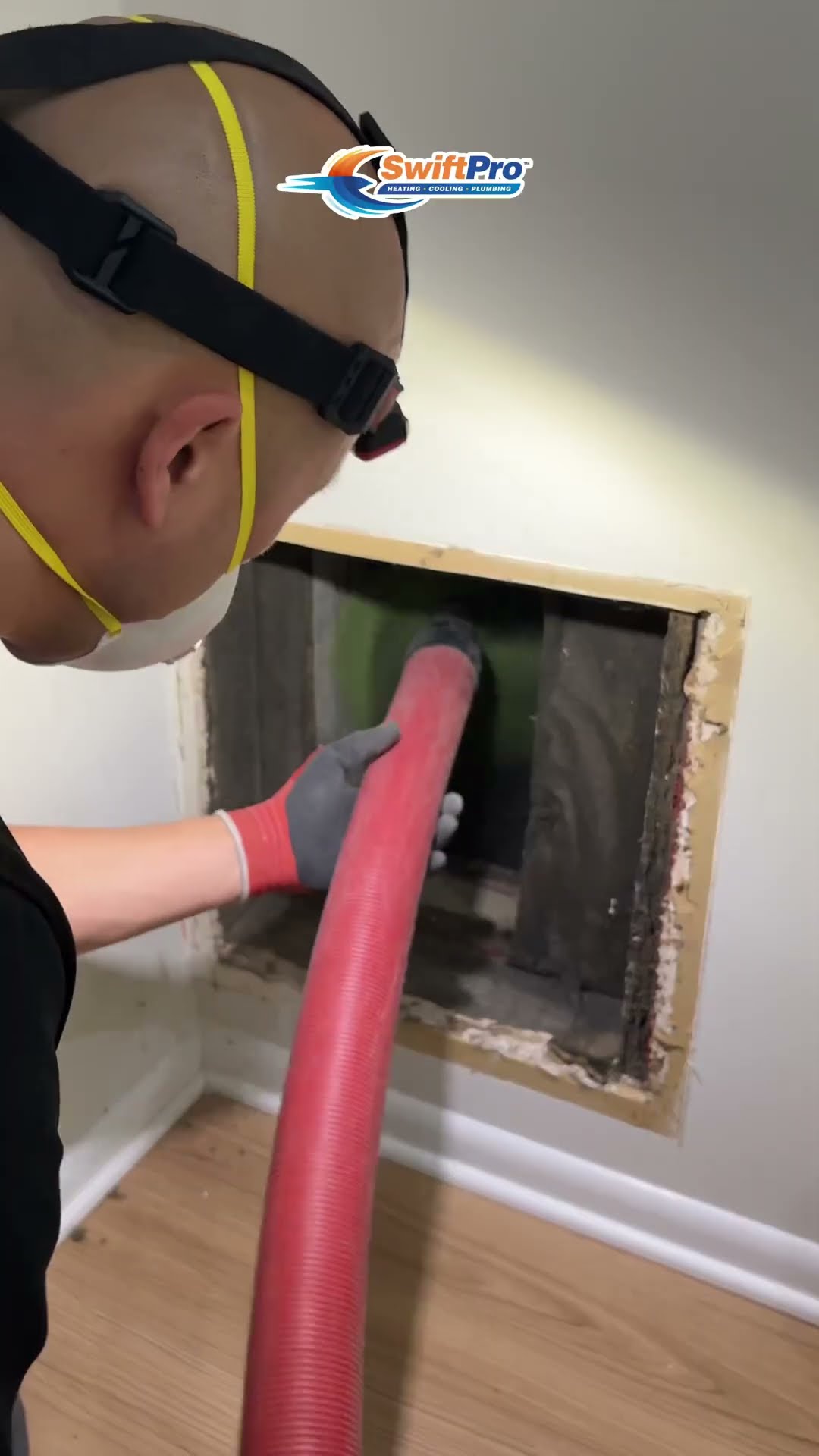 Duct cleaning: a tech in a headlamp and N95 mask runs a red vacuum hose into an open wall return in a home, kneels at a floor-level return grille, and the dusty inside of the duct is shown.