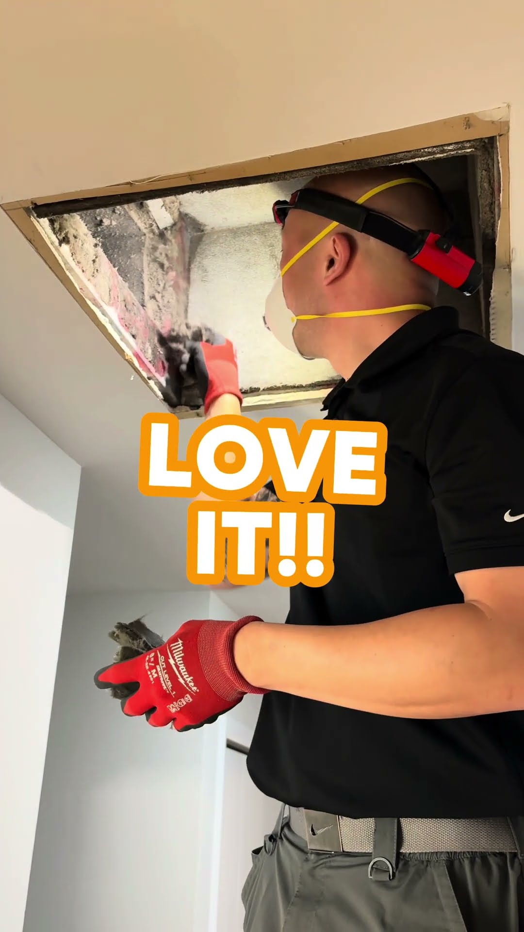 A duct cleaning tech in an N95 mask and red headlamp cleans an open ceiling duct opening, and vacuums a floor register in a carpeted room with a red hose.